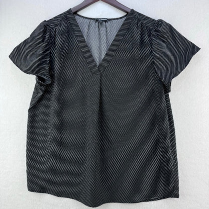 A black short-sleeve blouse with a subtle white polka dot pattern, featuring a V-neckline and puff sleeves, hanging on a dark wooden hanger against a light gray textured wall.