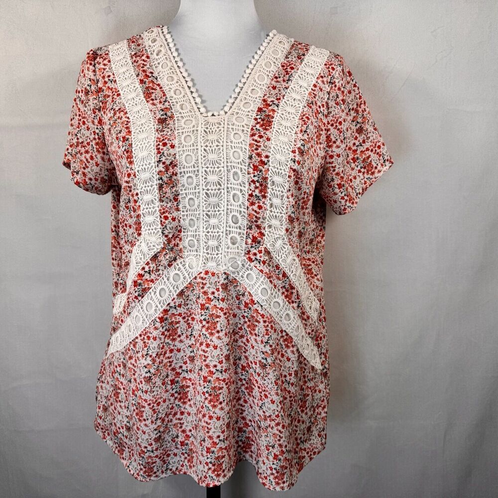 A short-sleeve blouse with a red and white floral print, featuring prominent white crochet lace panels down the front and along the V-neckline, displayed on a mannequin torso against a gray fabric backdrop.