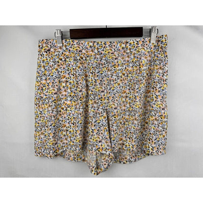 A pair of women's shorts featuring a dense, small-scale floral print in yellow, orange, blue, and white on a light gray base, hanging from a dark wooden hanger with silver metal clips against a light gray background.