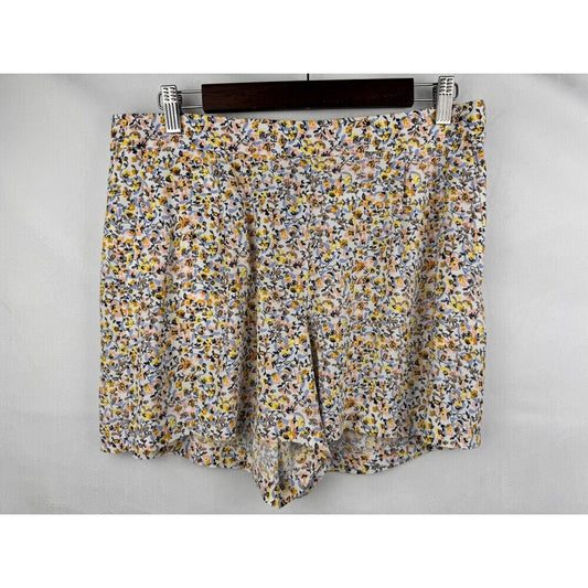 A pair of women's shorts featuring a dense, small-scale floral print in yellow, orange, blue, and white on a light gray base, hanging from a dark wooden hanger with silver metal clips against a light gray background.