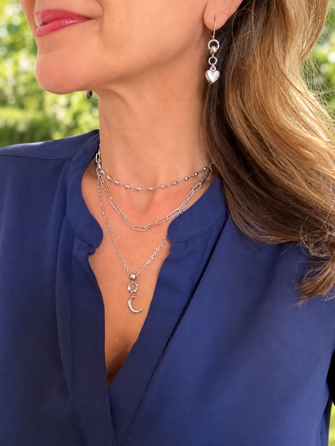 A close-up lifestyle shot of a woman wearing layered silver jewelry, including a silver crescent moon pendant necklace, a silver beaded choker, and a silver dangle earring with a heart charm, paired with a blue blouse.