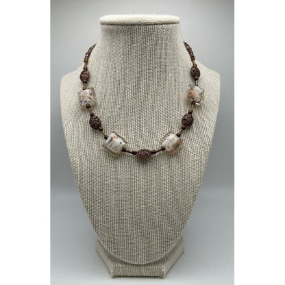 A beaded necklace displayed on a textured beige fabric jewelry bust. The necklace features white and beige marbled beads separated by dark brown textured spacer beads on a dark cord.