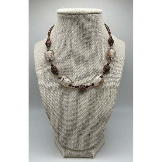 A beaded necklace displayed on a textured beige fabric jewelry bust. The necklace features white and beige marbled beads separated by dark brown textured spacer beads on a dark cord.