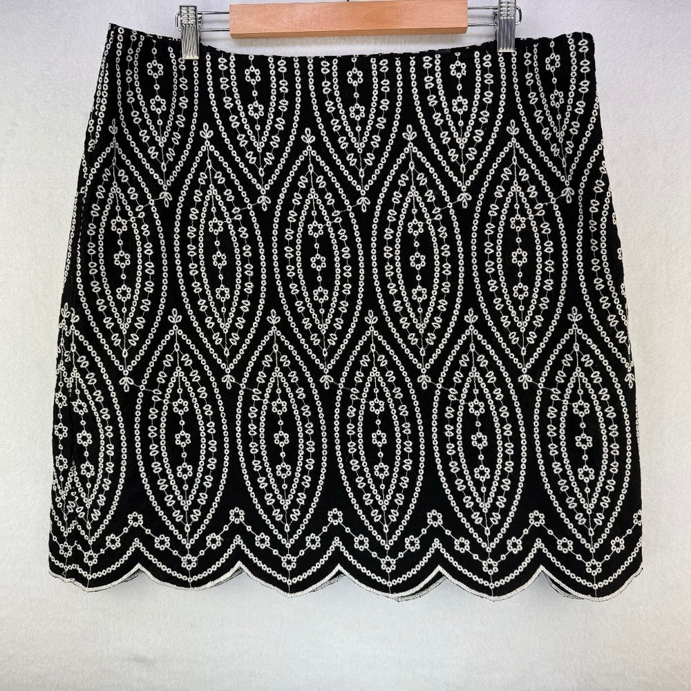 A black mini skirt with an intricate white geometric and floral pattern, featuring a scalloped hem and shown hanging on a wooden hanger against a white wall.