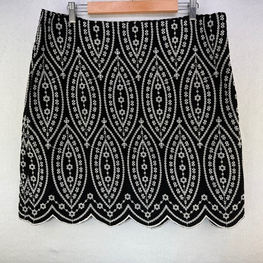 A black mini skirt with an intricate white geometric and floral pattern, featuring a scalloped hem and shown hanging on a wooden hanger against a white wall.