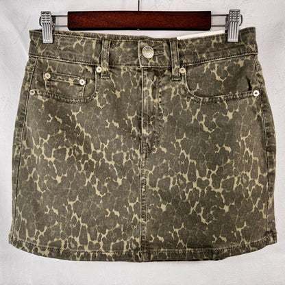 A vintage-style acid-wash denim mini skirt with a leopard print pattern, hanging on a wooden hanger against a light gray background.