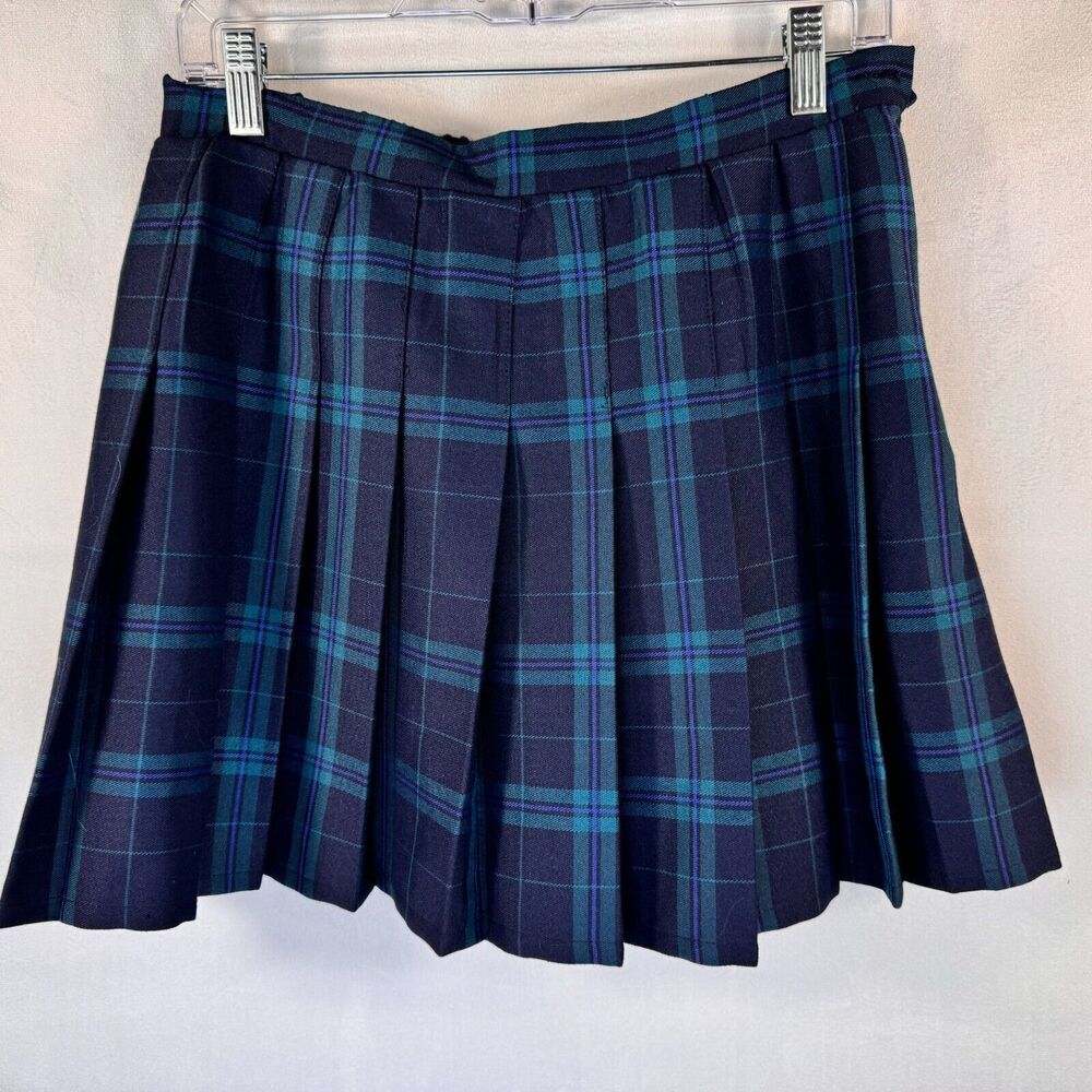 A pleated plaid mini skirt in navy blue with teal and light blue stripes, hanging from a clear plastic hanger against a light textured wall.