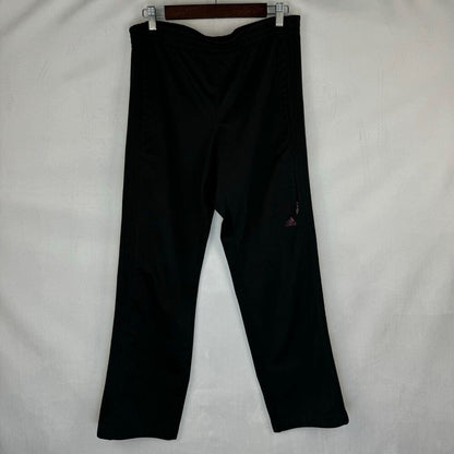 Black athletic pants hanging from a wooden hanger against a white fabric background, featuring a small red Adidas logo on the left leg.