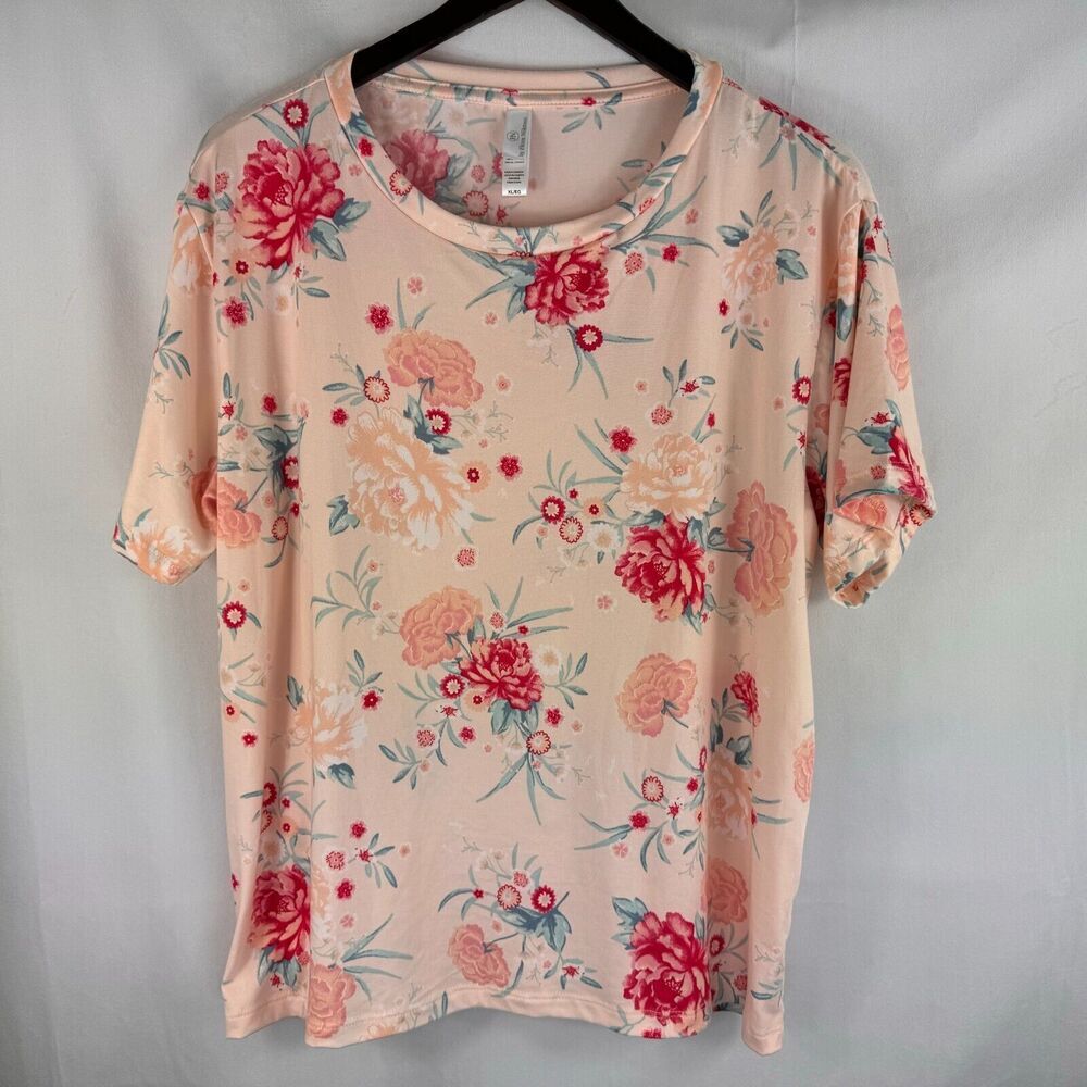 A short-sleeve crewneck blouse with a pinkish-beige base and a red, pink, and green floral print, hanging on a black hanger against a light gray backdrop.