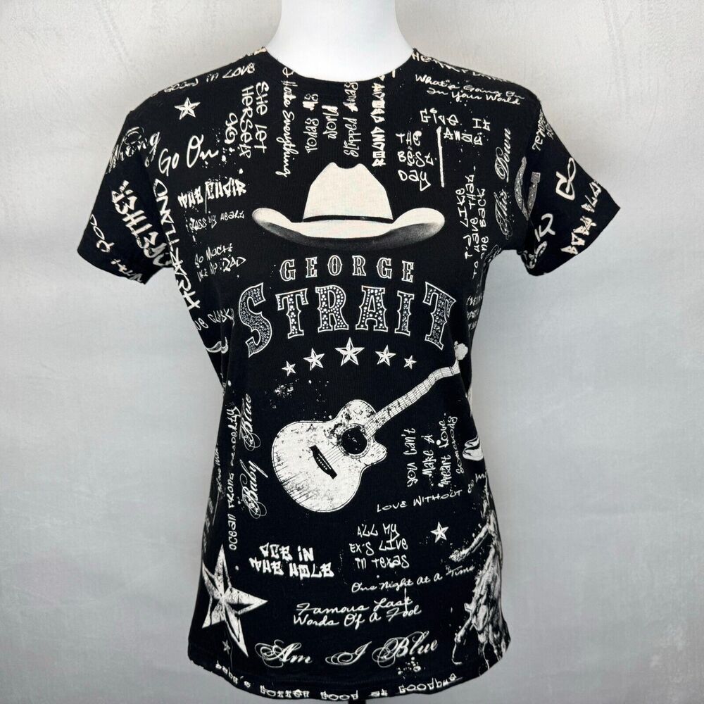 A black short-sleeve t-shirt displayed on a white mannequin torso against a light gray background. The shirt features a white graphic print with a cowboy hat, a guitar, and the text 'GEORGE STRAIGH' surrounded by handwritten-style text and stars.