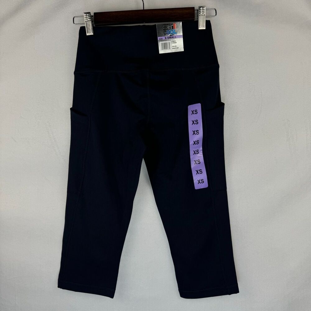 A pair of dark navy blue high-waisted capri leggings with side pockets, hanging on a wooden hanger against a light gray fabric background. A purple size tag is attached to the right leg.