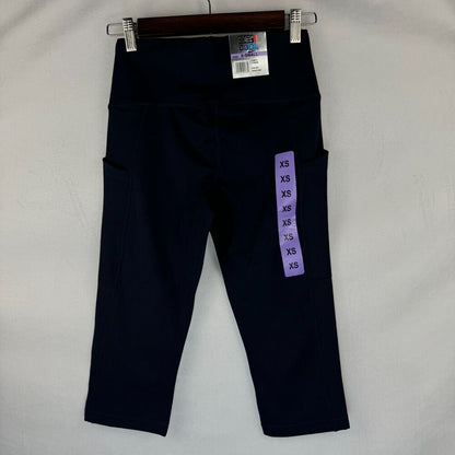 A pair of dark navy blue high-waisted capri leggings with side pockets, hanging on a wooden hanger against a light gray fabric background. A purple size tag is attached to the right leg.