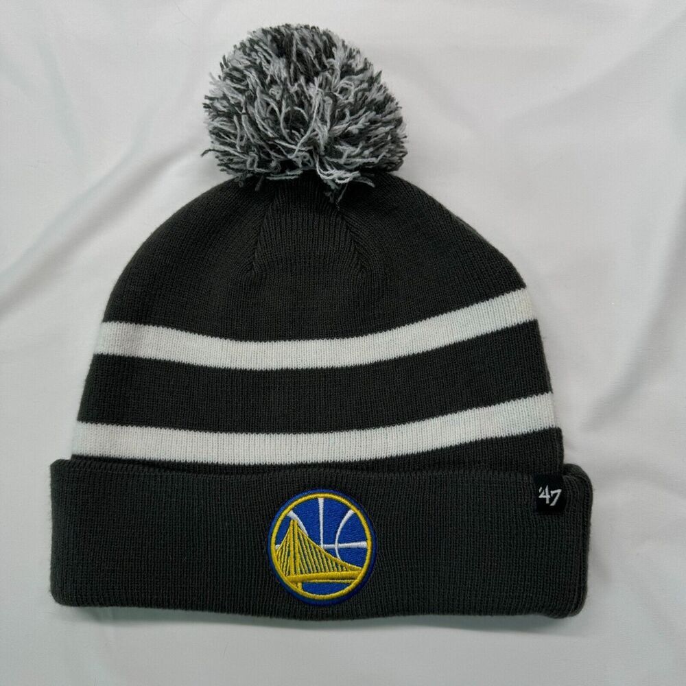 A black knit pom-pom beanie with two white horizontal stripes, featuring a Golden State Warriors logo patch on the front cuff and a '47 brand tag on the side, displayed on a white fabric background.
