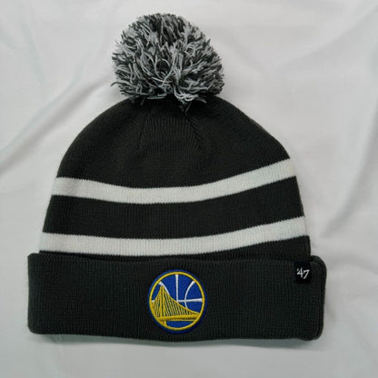 A black knit pom-pom beanie with two white horizontal stripes, featuring a Golden State Warriors logo patch on the front cuff and a '47 brand tag on the side, displayed on a white fabric background.