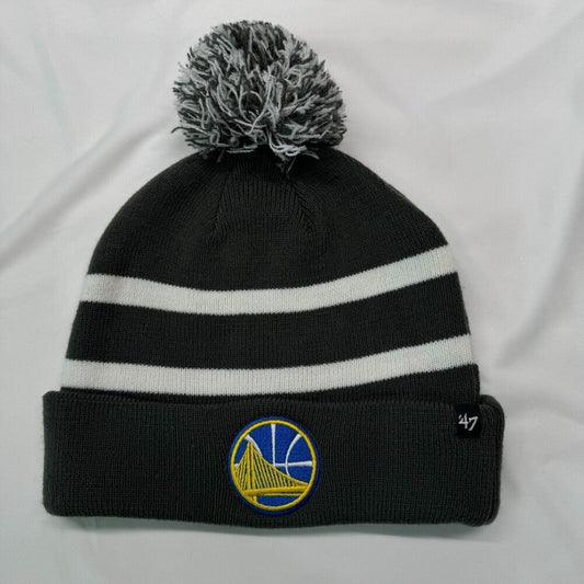 A black knit pom-pom beanie with two white horizontal stripes, featuring a Golden State Warriors logo patch on the front cuff and a '47 brand tag on the side, displayed on a white fabric background.