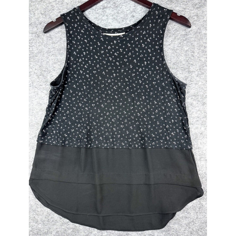 A sleeveless black top with a small white floral pattern on the upper half and a solid black ruffled overlay on the lower half, hanging on a dark wooden hanger against a gray textured background.