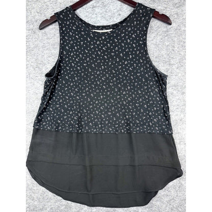 A sleeveless black top with a small white floral pattern on the upper half and a solid black ruffled overlay on the lower half, hanging on a dark wooden hanger against a gray textured background.