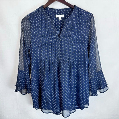 A navy blue blouse with a white geometric pattern, featuring sheer mesh bell sleeves and a V-neckline, hanging on a wooden hanger against a white background.