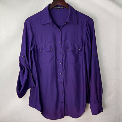 A purple long-sleeve button-up shirt with two chest pockets and rolled cuffs, hanging on a wooden hanger against a light gray fabric backdrop.