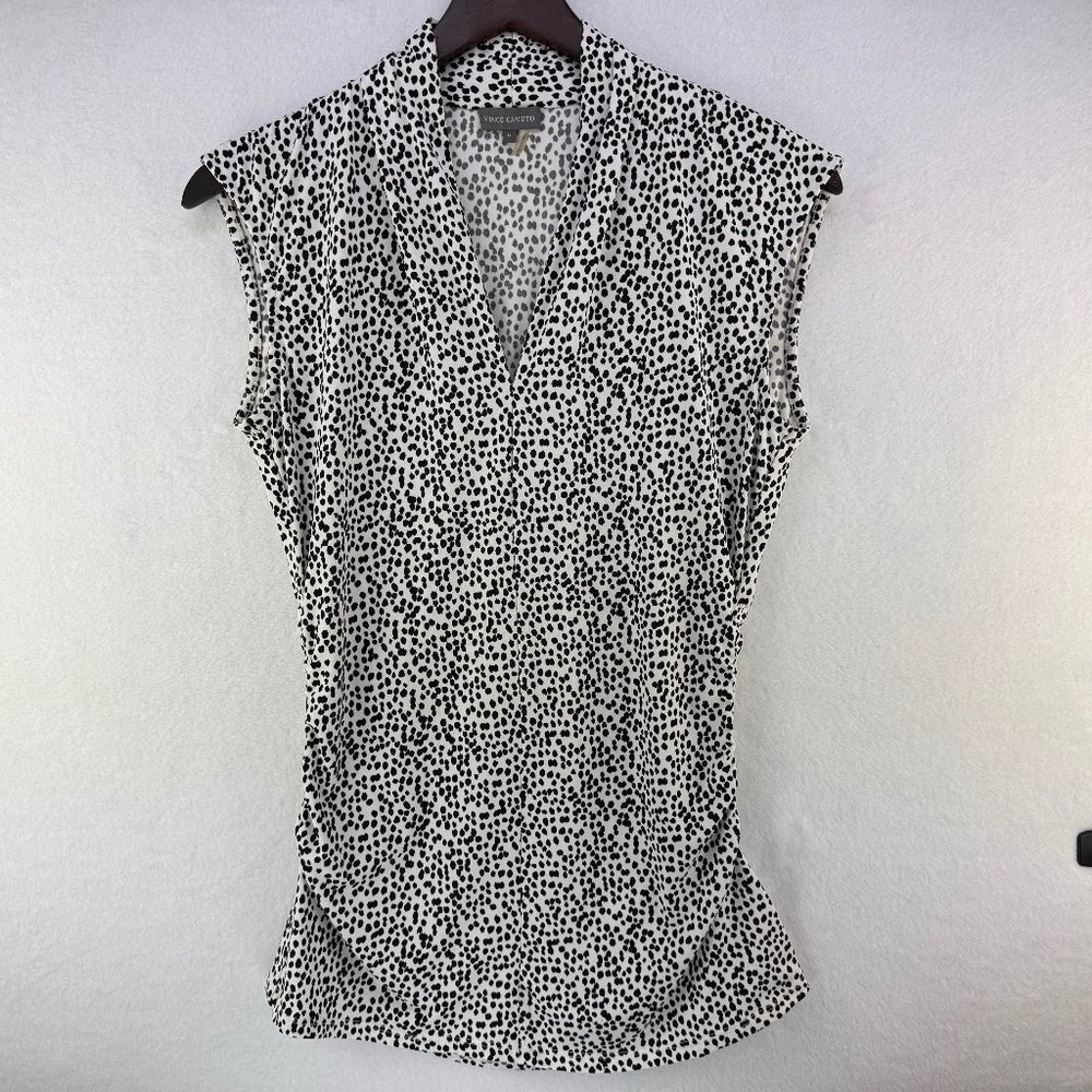 A sleeveless black and white spotted pattern blouse hanging on a dark hanger against a textured white wall.