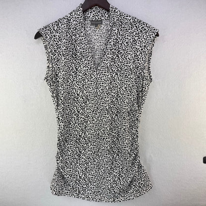 A sleeveless black and white spotted pattern blouse hanging on a dark hanger against a textured white wall.