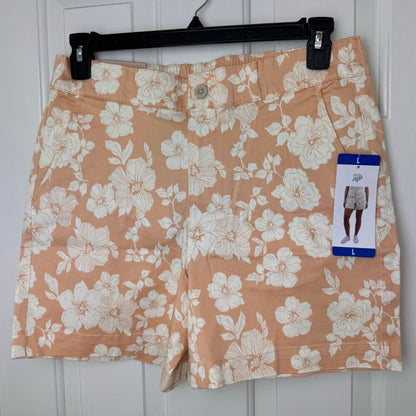 A pair of women's shorts in a peach-orange color with a white floral print, hanging on a black hanger against a white paneled door. The shorts still have their retail tag attached.