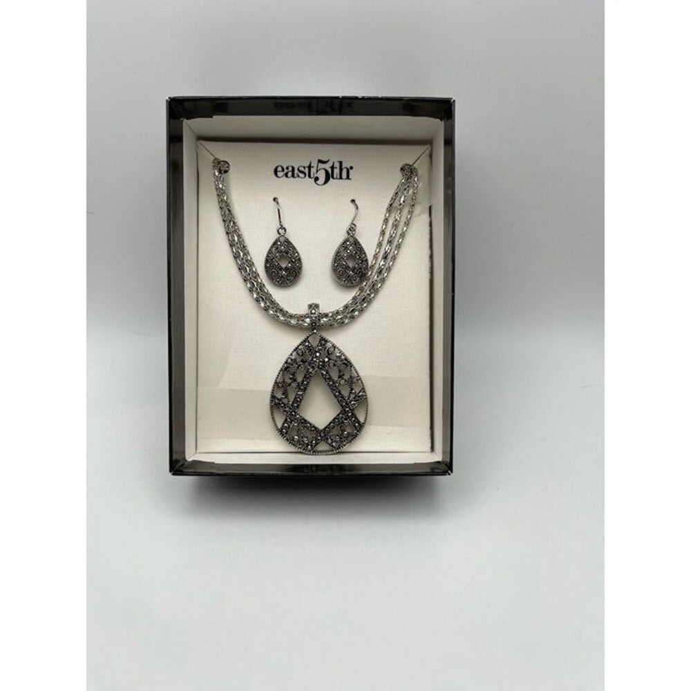 A silver-toned jewelry set featuring a teardrop pendant necklace and matching drop earrings, presented inside a black box with a cream insert. The brand name 'east5dr' is printed on the insert.