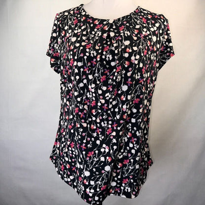 A black short-sleeve blouse with a pink, white,  floral print, displayed on a white mannequin torso against a light gray fabric backdrop.