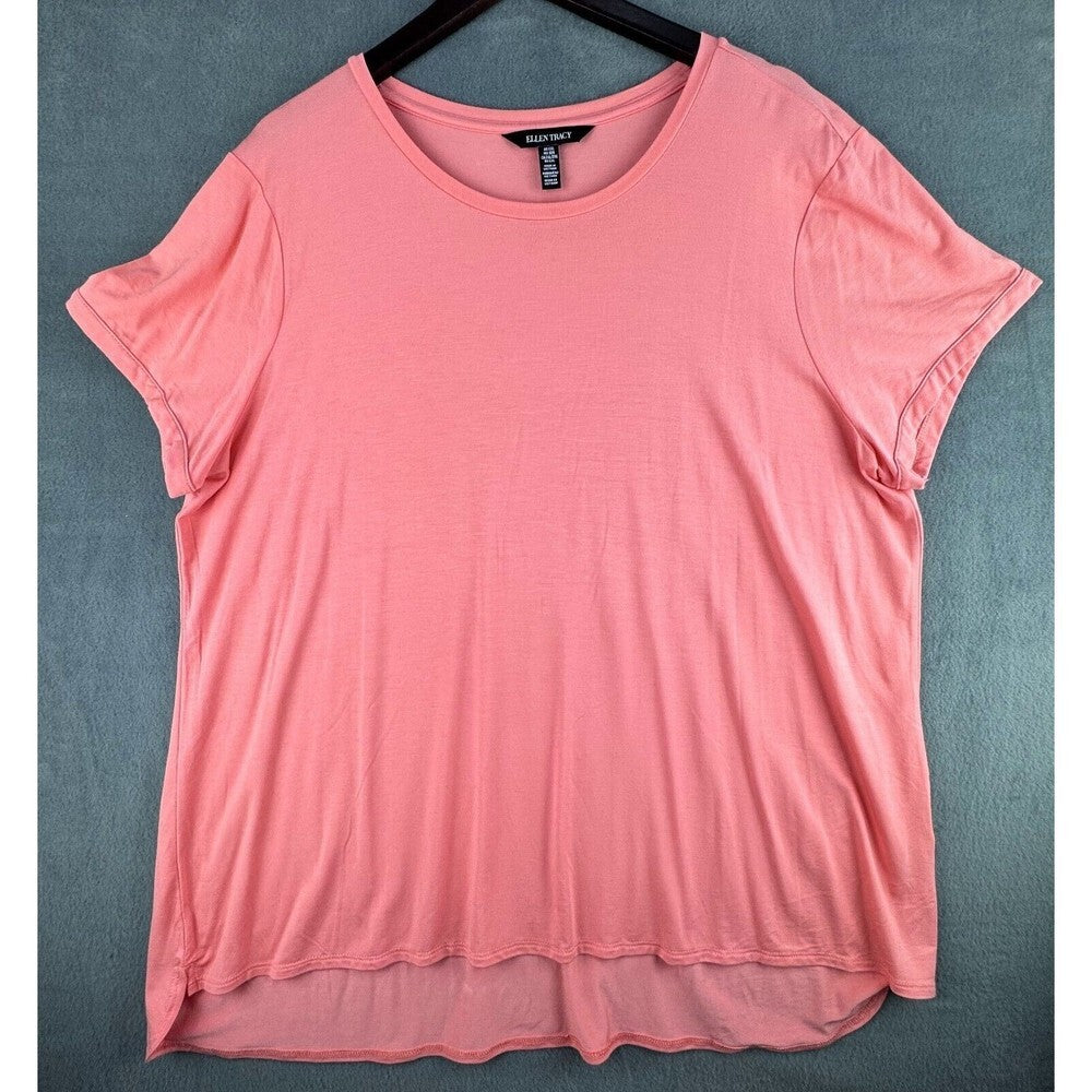 A coral pink short-sleeve crewneck t-shirt hanging on a black hanger against a textured gray background.