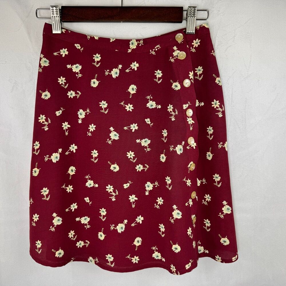 A burgundy A-line skirt with a small cream floral print, hanging on a dark wooden hanger against a textured white wall.