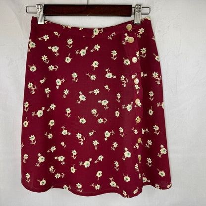 A burgundy A-line skirt with a small cream floral print, hanging on a dark wooden hanger against a textured white wall.