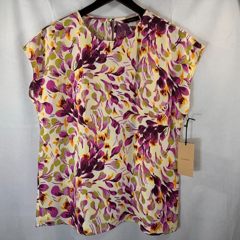 A short-sleeve blouse with a vibrant floral print in purple, green, and orange on a cream background, hanging on a black hanger with a paper tag attached.