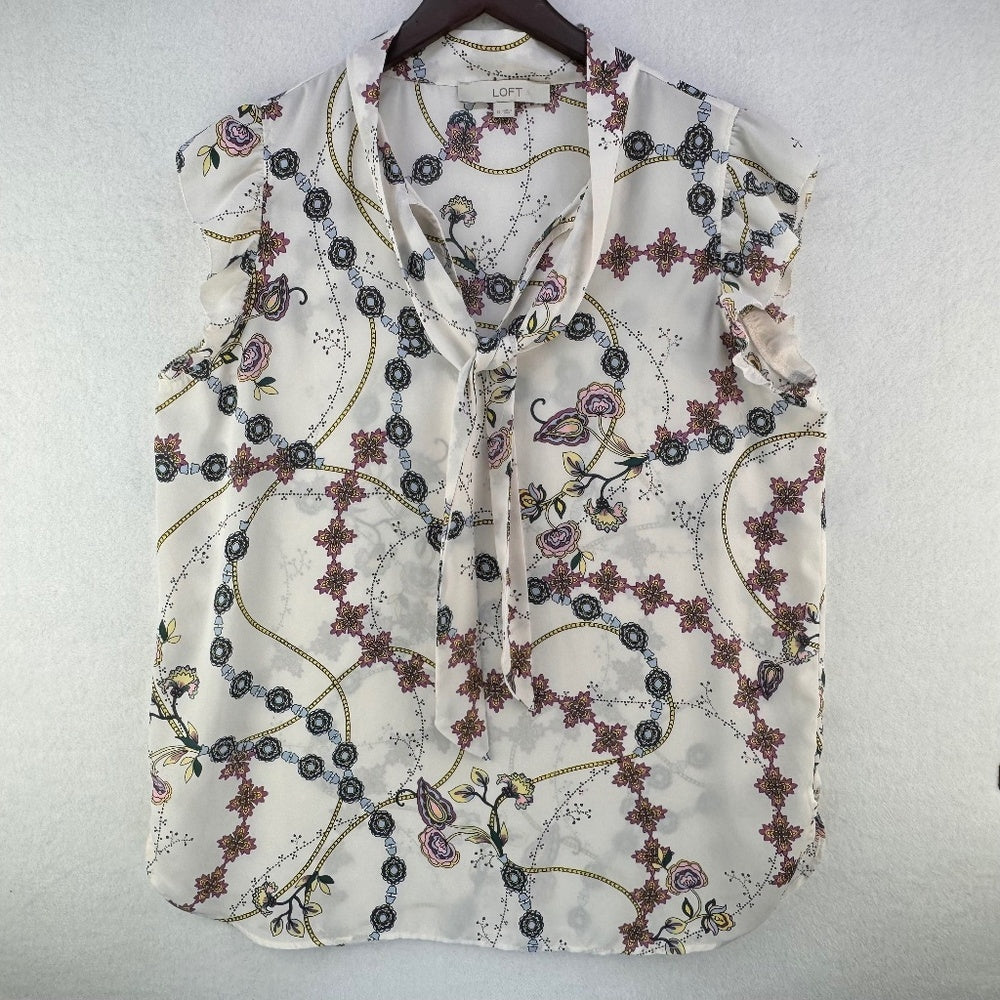 A sleeveless cream-colored blouse with a tie-neck and a delicate floral and vine pattern, hanging on a dark wooden hanger against a textured white wall.