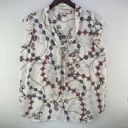 A sleeveless cream-colored blouse with a tie-neck and a delicate floral and vine pattern, hanging on a dark wooden hanger against a textured white wall.