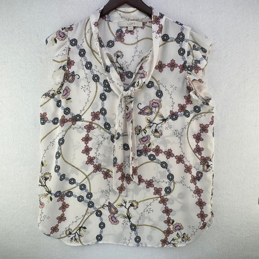 A sleeveless cream-colored blouse with a tie-neck and a delicate floral and vine pattern, hanging on a dark wooden hanger against a textured white wall.