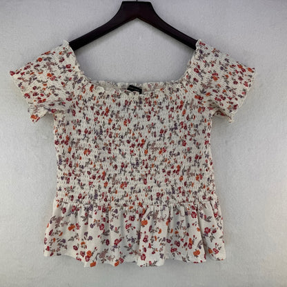 A women's short-sleeve smocked blouse with a ruffled hem and off-the-shoulder neckline, featuring a red, orange, and purple floral print on a cream background, hanging on a dark wooden hanger against a light gray textured wall.