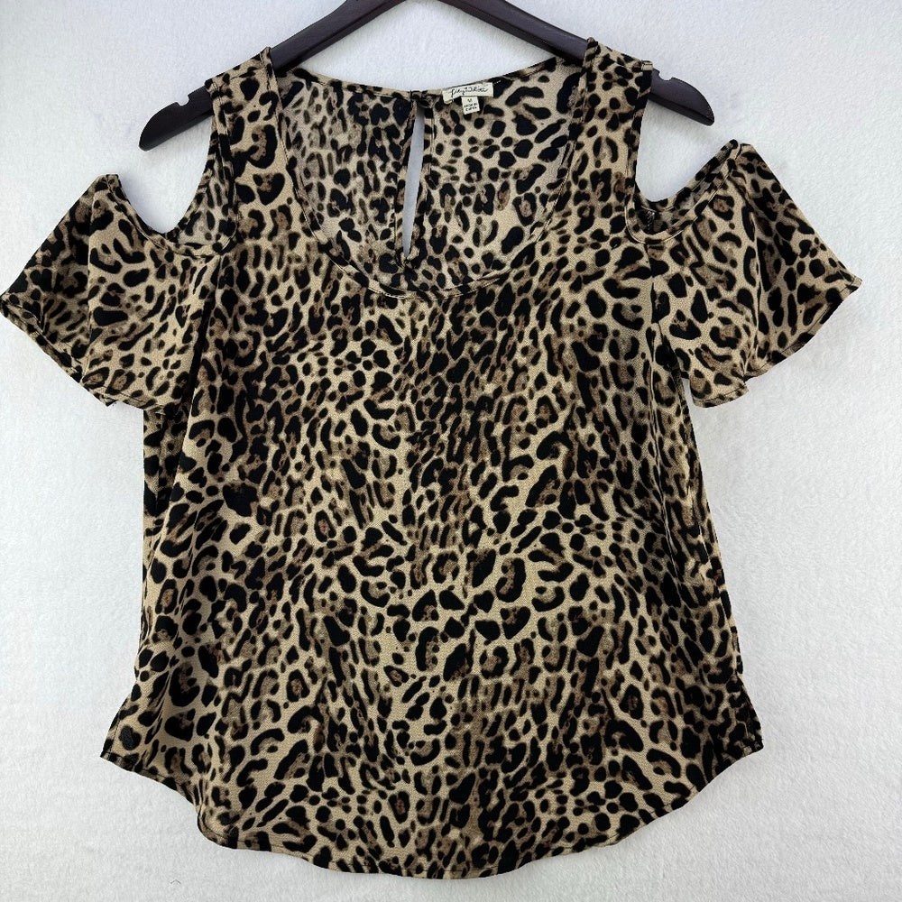A leopard print cold-shoulder top hanging on a black hanger against a white textured wall. The top features a beige and brown base with black leopard spots and short puff sleeves.