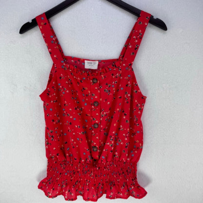 A red sleeveless top with a small multicolor floral print, wide shoulder straps, a button-up front, and an elasticized ruffled hem, hanging on a black hanger against a white textured wall.