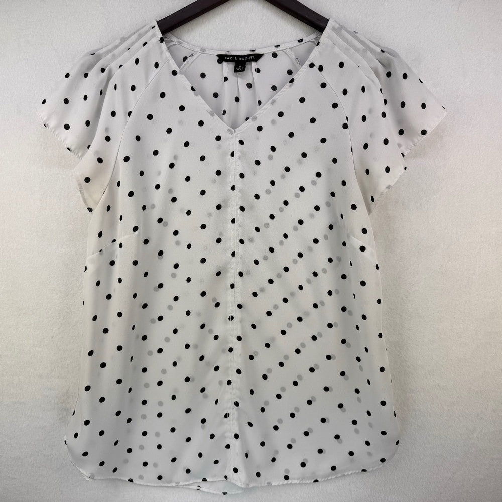 A white short-sleeve blouse with black polka dots, featuring a V-neckline and shoulder cutouts, hanging on a black hanger against a textured white wall.