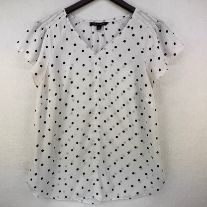 A white short-sleeve blouse with black polka dots, featuring a V-neckline and shoulder cutouts, hanging on a black hanger against a textured white wall.