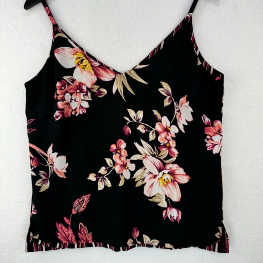 A black camisole tank top with a vibrant floral print in pink, white, and beige, featuring thin spaghetti straps and a ruffled hem, displayed on a white textured background.
