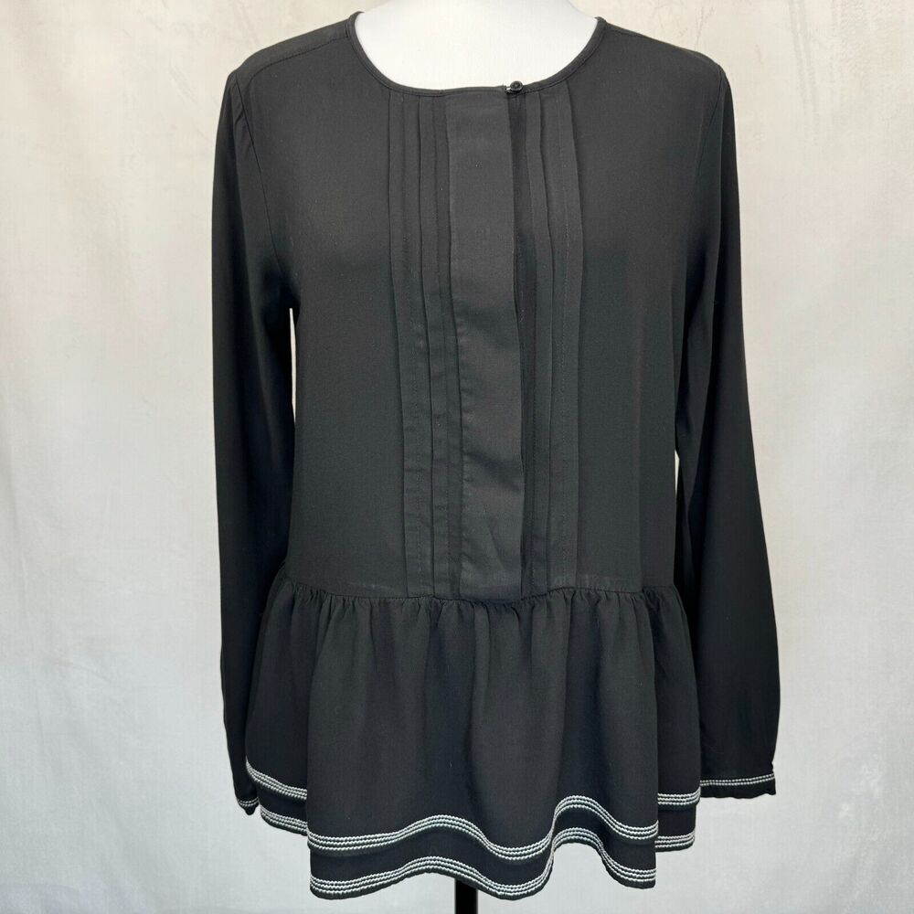 A black long-sleeve blouse with vertical pleats on the front and a ruffled hem, displayed on a mannequin against a light gray fabric backdrop.