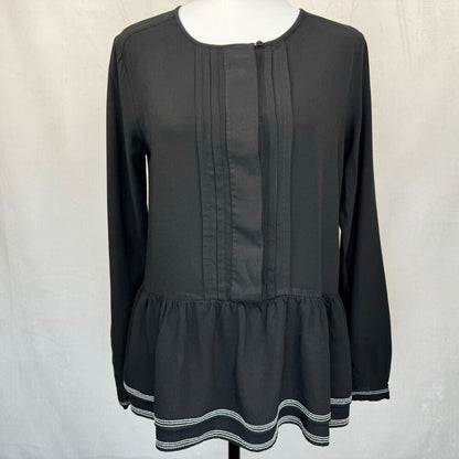 A black long-sleeve blouse with vertical pleats on the front and a ruffled hem, displayed on a mannequin against a light gray fabric backdrop.