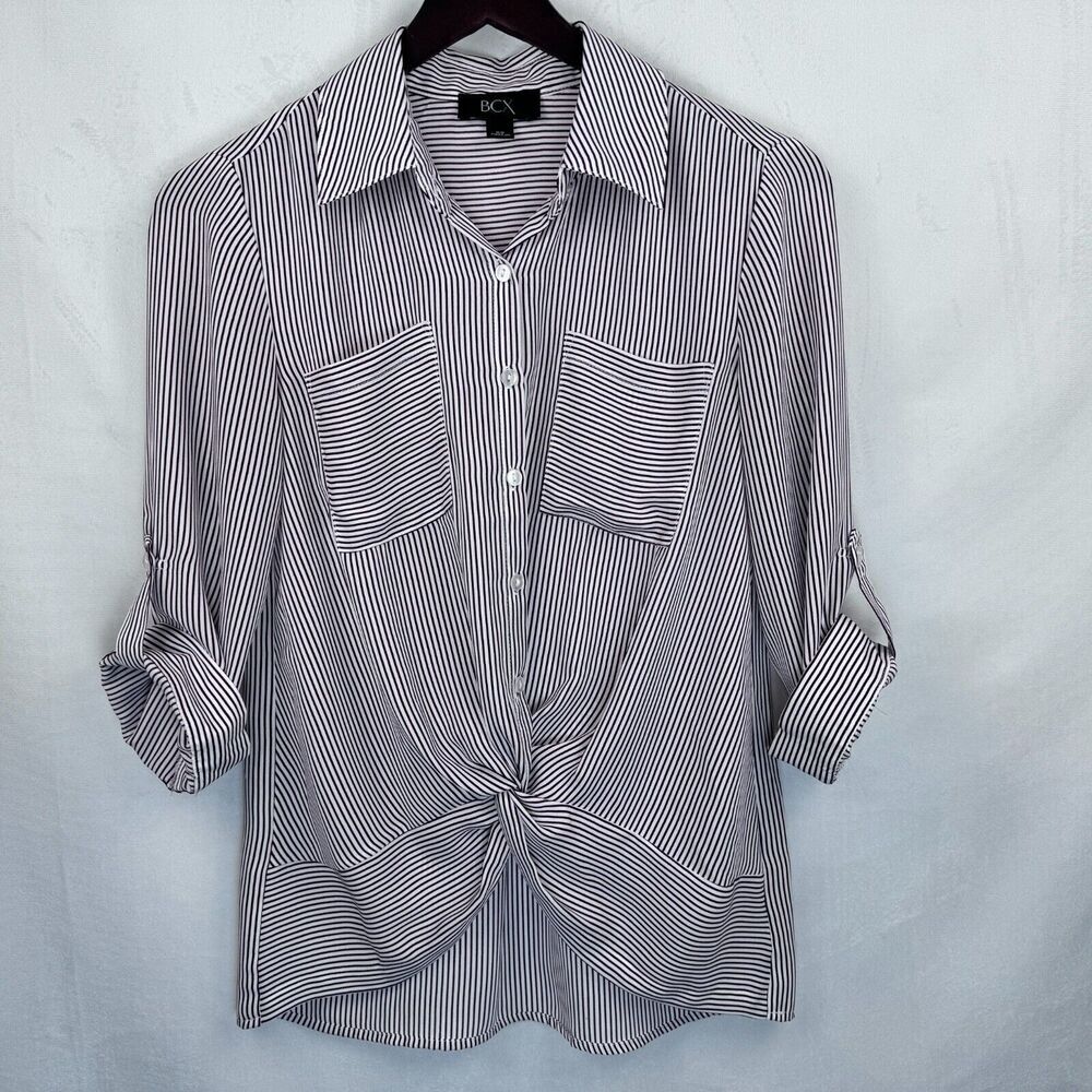 A women's long-sleeve button-up shirt with thin vertical black and white stripes, featuring two chest pockets and a front knot detail, hanging on a black hanger against a light gray textured backdrop.
