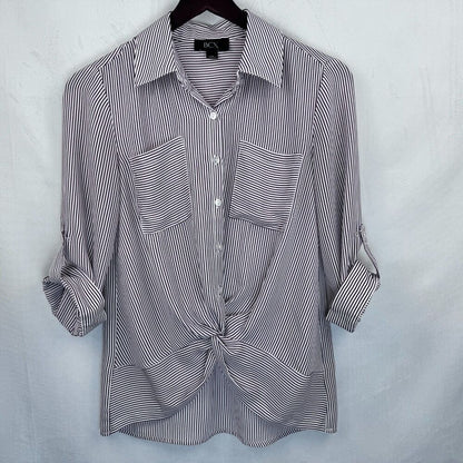 A women's long-sleeve button-up shirt with thin vertical black and white stripes, featuring two chest pockets and a front knot detail, hanging on a black hanger against a light gray textured backdrop.