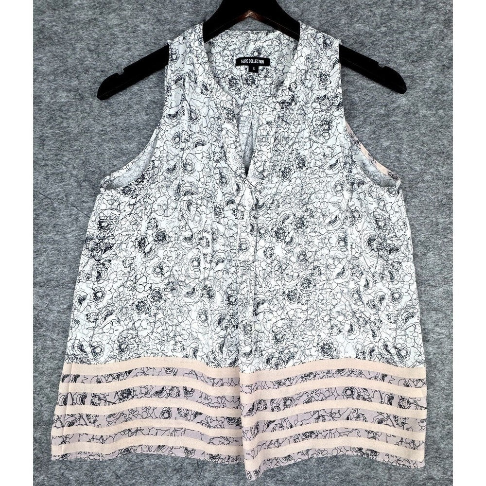 A sleeveless white top with a black floral pattern and a tiered hem featuring beige stripes, hanging on a black hanger against a gray textured background.