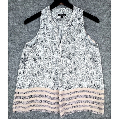 A sleeveless white top with a black floral pattern and a tiered hem featuring beige stripes, hanging on a black hanger against a gray textured background.
