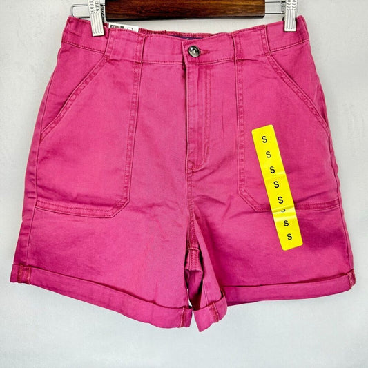 Bright pink denim shorts with a yellow size tag hanging from the side, displayed on a wooden hanger against a light gray wall.