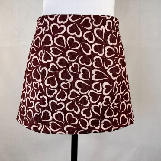 A high-waisted A-line mini skirt with an all-over repeating heart pattern in dark burgundy and cream, displayed on a white mannequin torso.