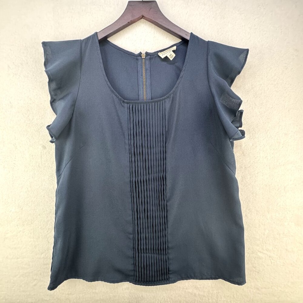 A slate blue sleeveless blouse with ruffled cap sleeves and a vertical pleated front panel, hanging on a wooden hanger against a textured beige wall.